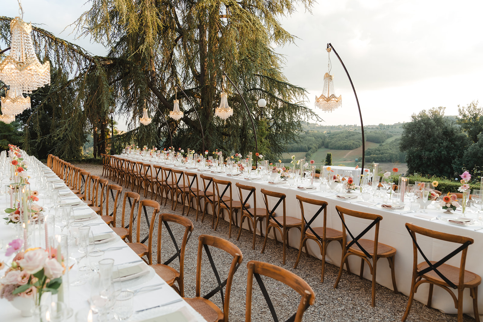 Long outdoor wedding table set under trees with chandeliers and Tuscan hills beyond